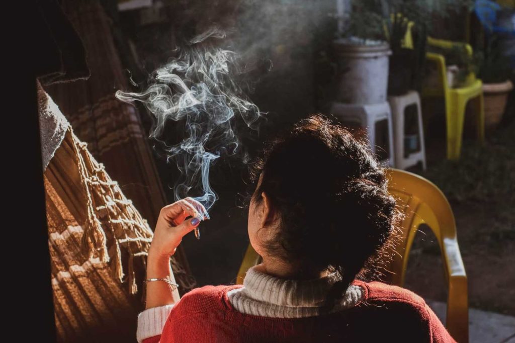 A woman in a red sweater smokes a joint with potted plants in the background.