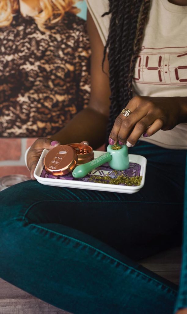 Black woman seated wearing jeans and holding a tray of cannabis products with a pipe